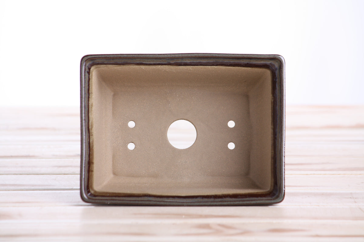 Rectangular ceramic bonsai pot with six drainage holes, light brown interior, dark rim, placed on wooden surface.