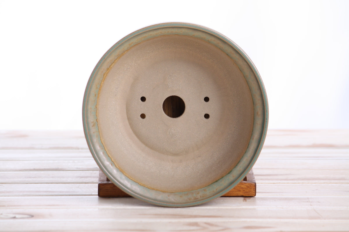 Bottom view of a round ceramic bonsai pot with a hole and speckled finish resting on a wooden stand on a light-colored wooden surface.