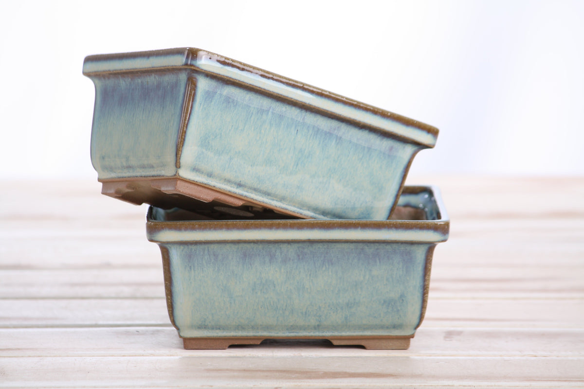 Two light blue ceramic bonsai pots with a frosty glaze stacked on a wooden surface, with natural light in the background