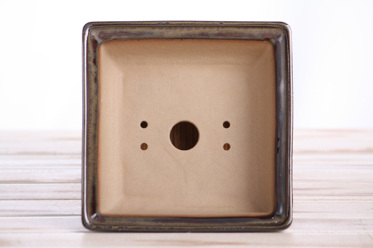 Bottom view of a square ceramic bonsai pot with dark honey glaze and drainage holes, placed on a light wooden surface.