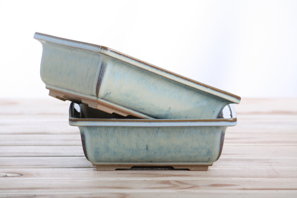 Stacked square ceramic bonsai pots with a Glacier glaze, placed on a wooden surface against a plain backdrop.