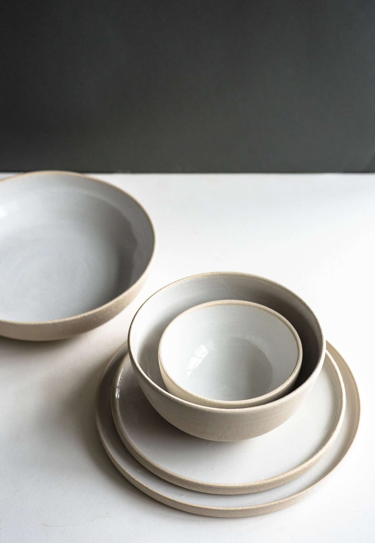 A handmade stoneware small bowl with a neutral finish, displayed alongside a stack of plates and a larger bowl, all set on a plain white surface against a dark backdrop.