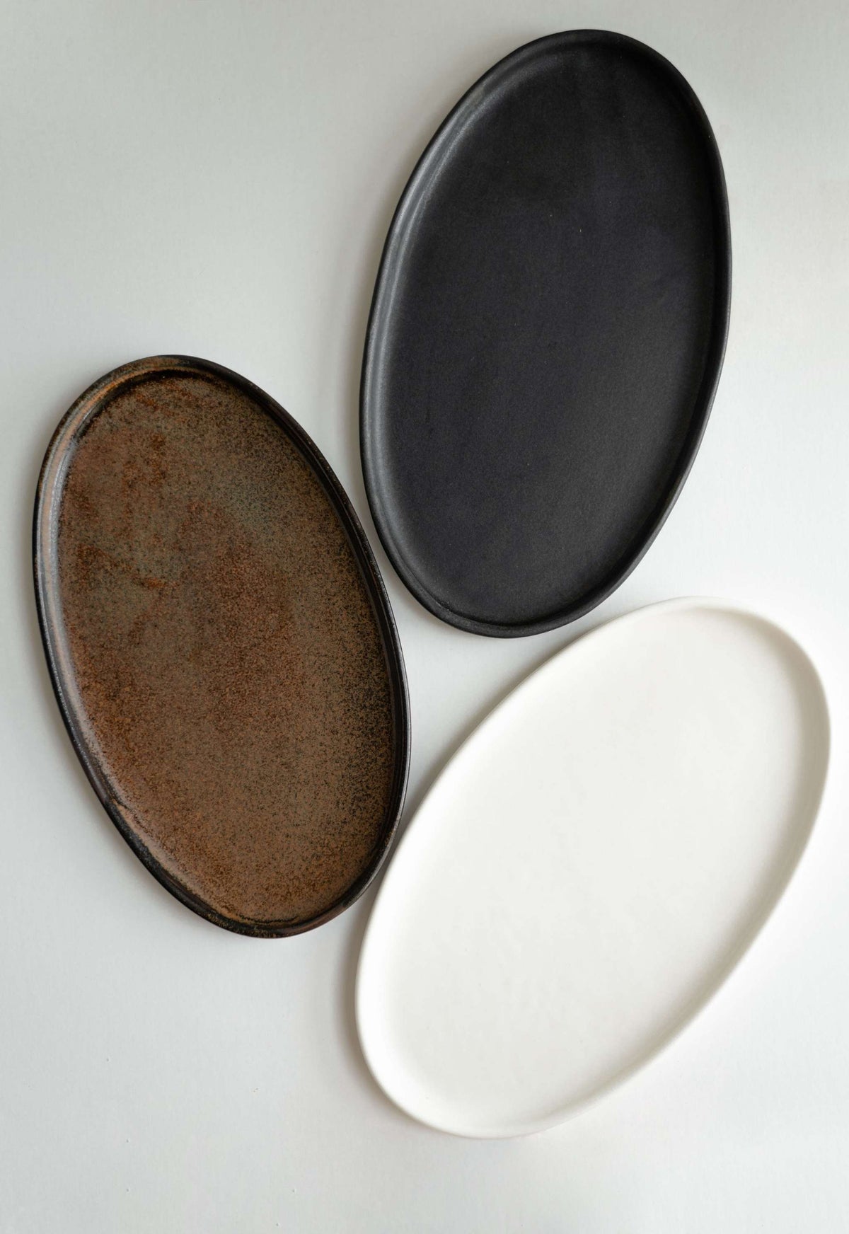 Three oval stone platters in rustic matte finishes, one dark brown, one black, and one white, on a plain surface.