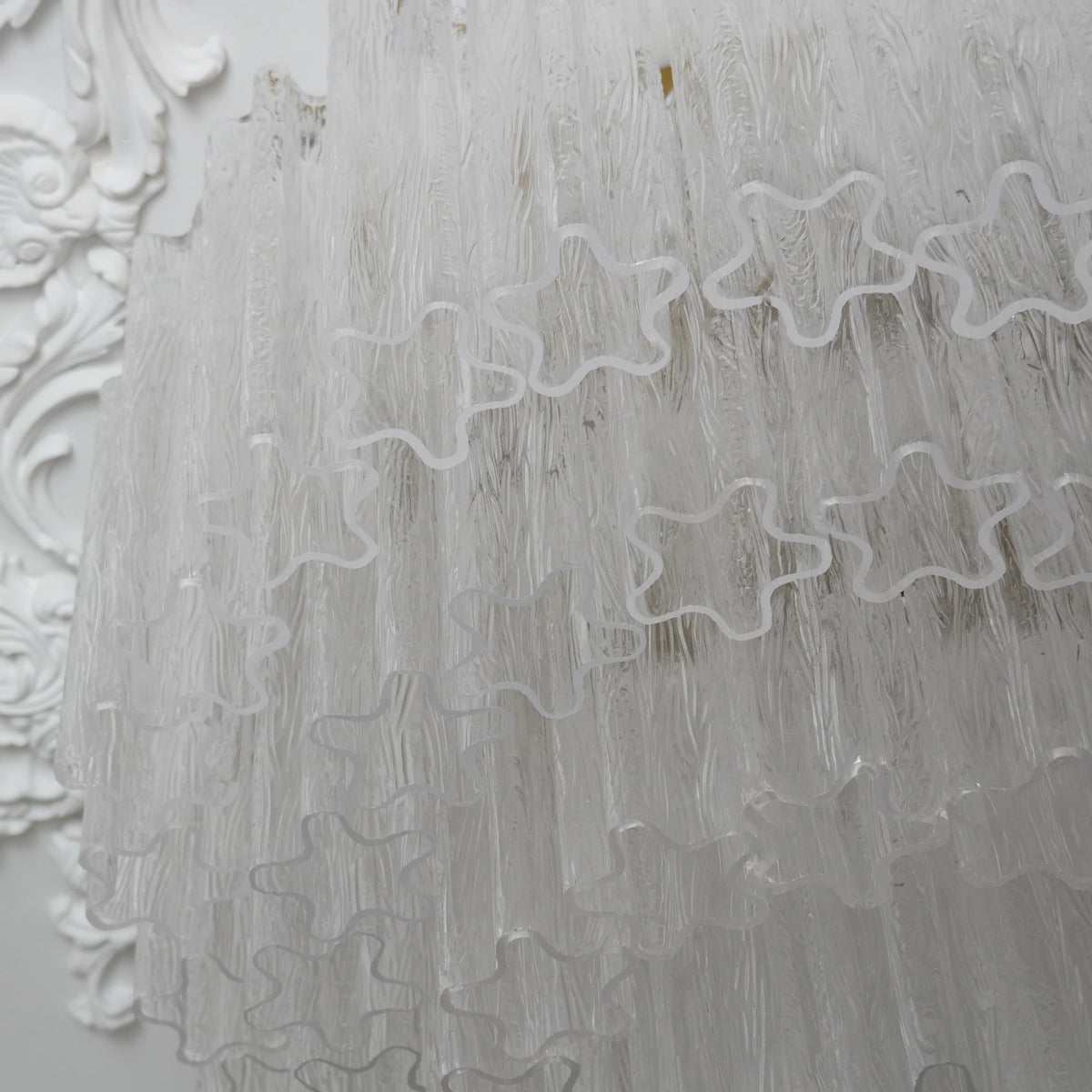 Clear, textured Murano glass chandelier with star cutouts, set against ornate white wall.