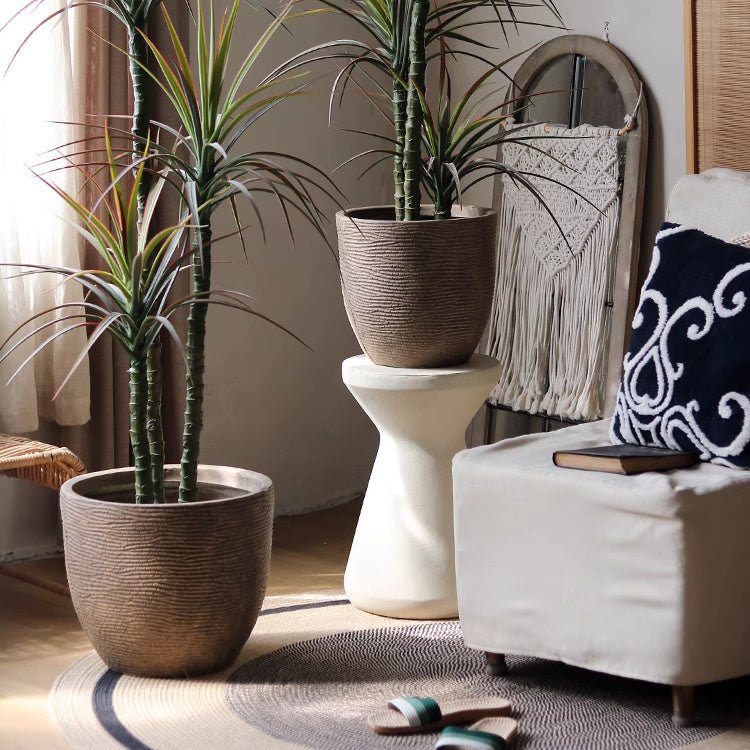 Two wooden planters with green plants, one on a stone pedestal, next to a white armchair and wooden sandals on a rug.