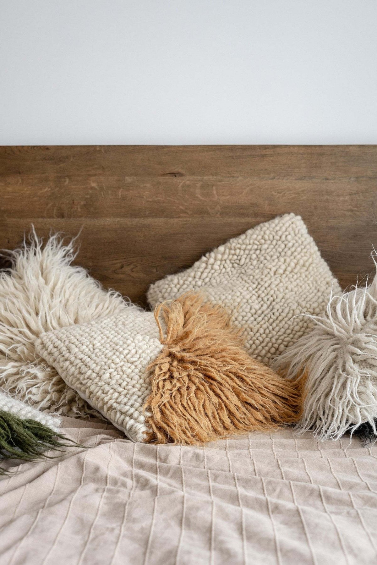A handwoven wool decorative pillow with fringe in a neutral color on a bed with a beige quilted blanket and wooden headboard.