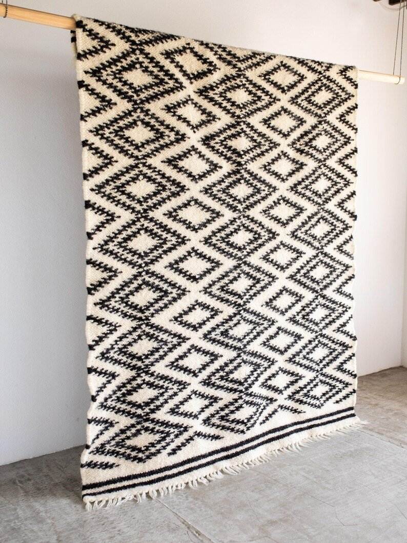 A black and white patterned rug hanging vertically on a wooden rod against a white wall, showing the traditional Carpathian Wool Lizhnyk - "Spectacled" Pattern.