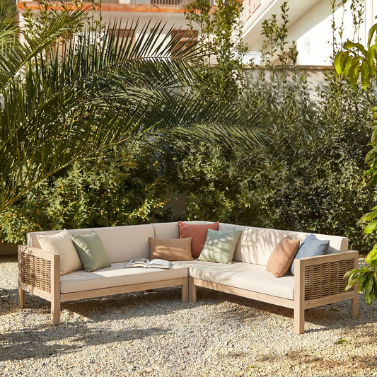 A beige woven sofa with colorful cushions, set outdoors on a gravel patio surrounded by greenery.