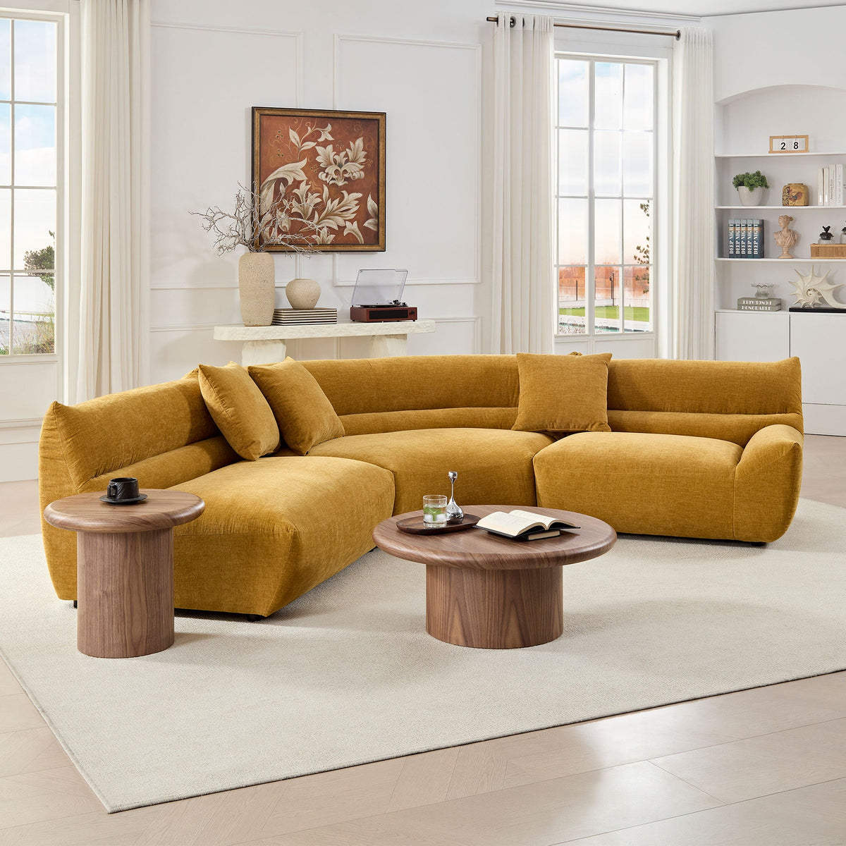 A mustard-colored modular curved sectional sofa paired with wooden side and coffee tables in a bright, minimalist room with a white rug and a wall with abstract art