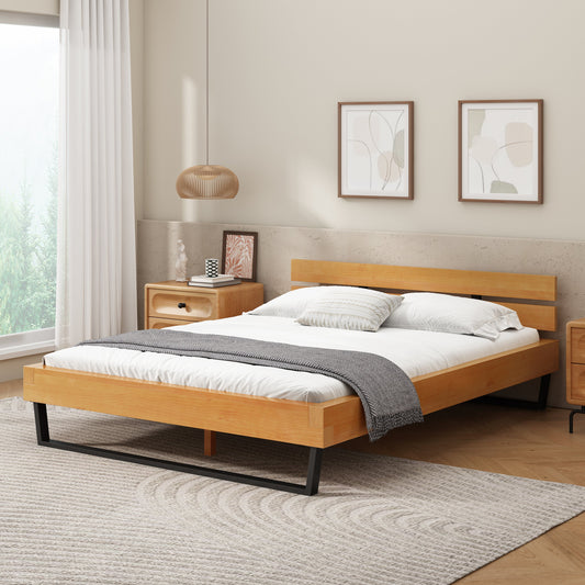 A queen size bed with a natural pine wood frame and black metal legs, neatly made with white bedding a dark grey throw, hanging light, white curtains, and framed artwork on a concrete wall.