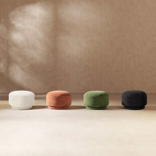 Four round footstools in white, terracotta, dark green, and black lined up on a neutral floor with a textured wall in the background.