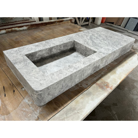A rectangular marble sink with a deep basin, showcasing natural veining and grey tones, placed on a wooden surface in a workshop setting.