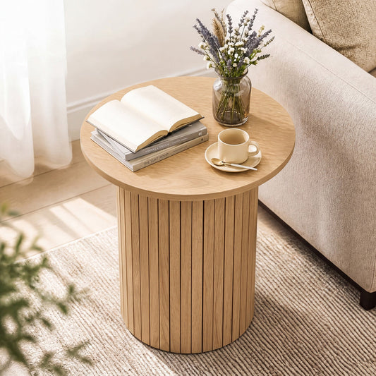 Round wooden side table with fluted legs, placed on a beige carpet near a window with white curtains. A coffee cup, saucer, book, and vase with dried flowers sit on the table.