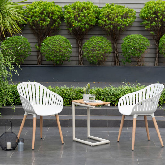 Two white C-shape chairs with light wooden legs arranged around a small table outdoors with greenery behind.