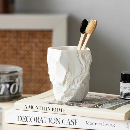 A textured white ceramic wind toothbrush cup containing black and yellow toothbrushes, placed on books in a light-colored room.