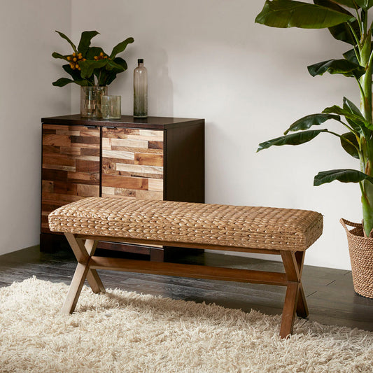 A woven water hyacinth bench with chestnut mahogany legs on a shag rug next to a wood dresser and a large potted plant