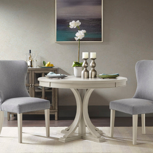 Round dining table with two chairs, antique cream wood veneer top, and solid wood pedestal legs in a light gray setting with decorative items and a painting on the wall.