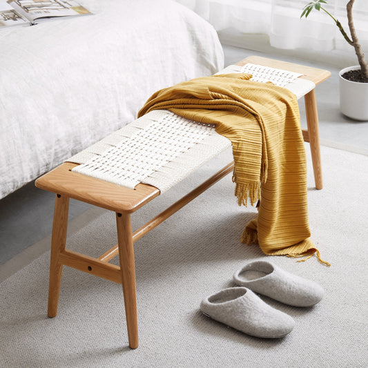 A Solid Wood Bench with a Handwoven Cane Seat, draped with a yellow knit throw, placed in a minimalist bedroom setting with slippers on the floor.