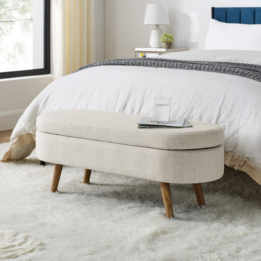 Oval storage bench with beige fabric and rubber wood legs, positioned on a fluffy white rug with a bed in the background.