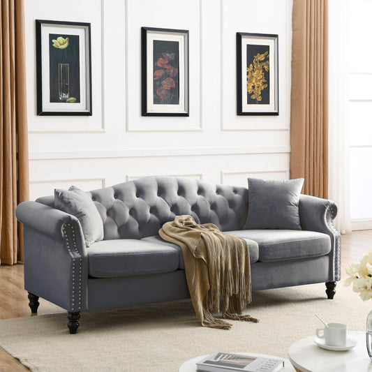 Grey Chesterfield sofa with button-tufted seat, adorned with a tan throw blanket and accompanied by beige pillows, set in a room with framed flower prints on the wall, light wood floors, and beige curtains.