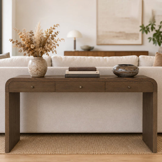 Rectangular brown wooden table with three drawers, placed in front of a beige sofa, with a vase and books on top.