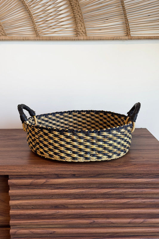 Circular tray with natural and black checkered pattern, made of woven material, displayed on a wooden table