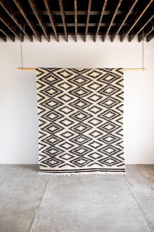 Wall hanging with geometric diamond patterns in black and white with fringes at the bottom, displayed on wooden supports against a plain wall.