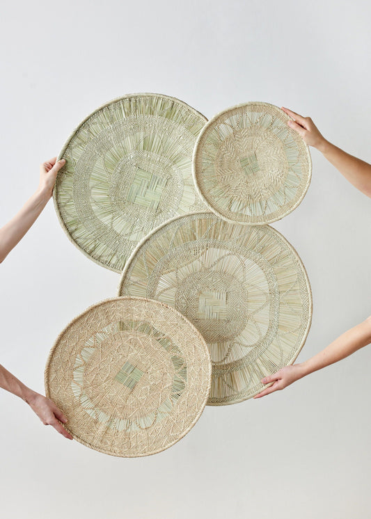 Four round baskets with intricate geometric patterns, held by two hands. Beige material with natural tones, plain background.