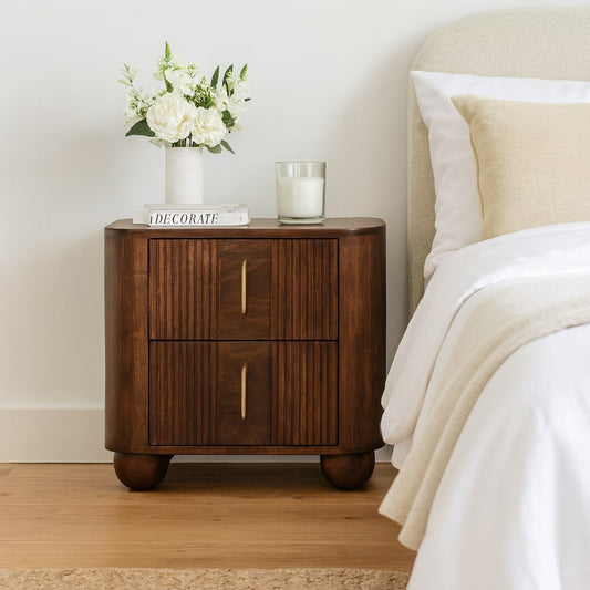 A wooden nightstand with two drawers and a vase of white flowers on top, placed next to a bed. Woodgrain finish, dark brown, near neutral wall.