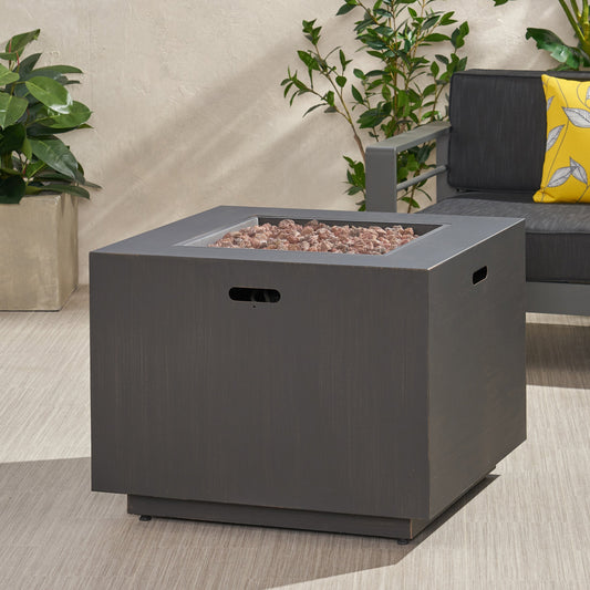 A square fire pit with a brushed brown finish, containing red and pink fire pits on a wooden deck with plants and seating in the background.