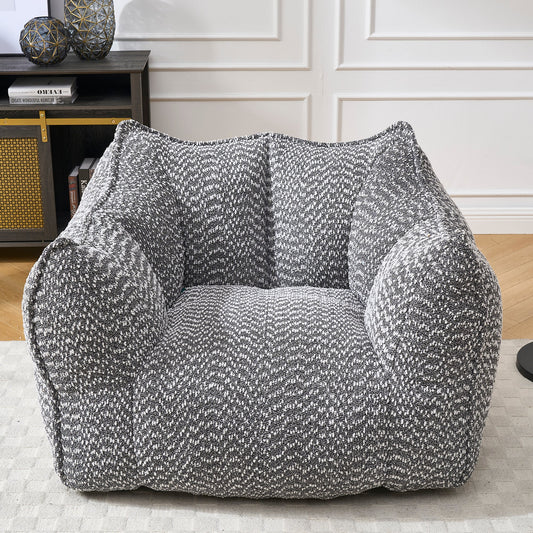 Gray and white speckled fabric square armchair on a white rug with a dark bookshelf and decorative items in the background