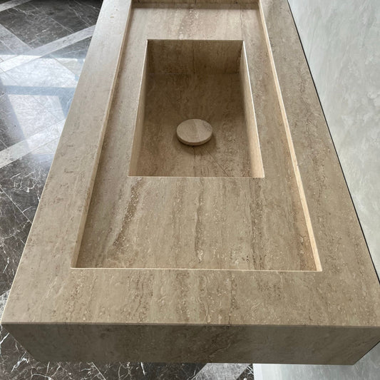 Rectangular travertine natural stone bathroom sink with a recessed basin and circular drain plug, placed on a tiled floor; setting is indoors with marble tiles in background.