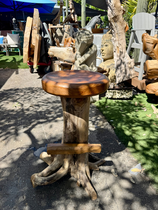 Handcrafted teak round bar stool with rough, unfinished wooden seat and legs placed outdoors under a blue umbrella, surrounded by sculptures and teak wood.