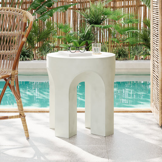Antique white round top side table with carved arches near a pool with plants and a wicker chair in the background