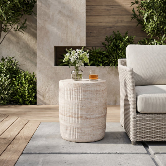 A fluted drum-shaped concrete side table with a simple floral arrangement and a glass of whiskey on top, placed outdoors with a wicker chair and greenery in the background.