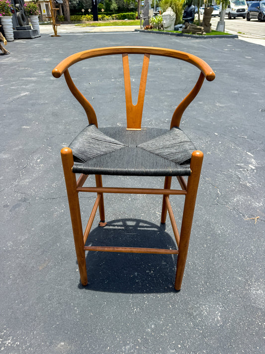 Wishbone Island Counter Stool. Light wood frame with woven seat, placed on pavement.