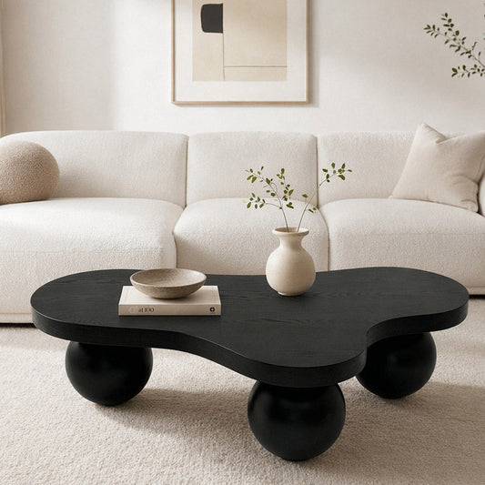 Black, organic-shaped coffee table with spherical legs, placed in a minimalist living room setting. A vase and a bowl sit on the table.
