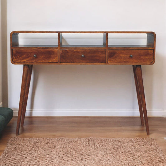 A solid wood triple curved chestnut console table with three drawers and a glass shelf on top, placed on a wooden floor with a rug in front.