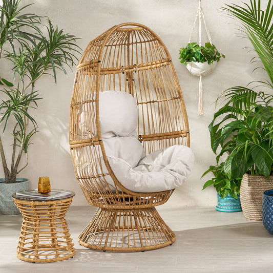 Rattan egg chair with cushioned seat and backrest, light brown color, accompanied by a small wicker side table; potted plants in the background.