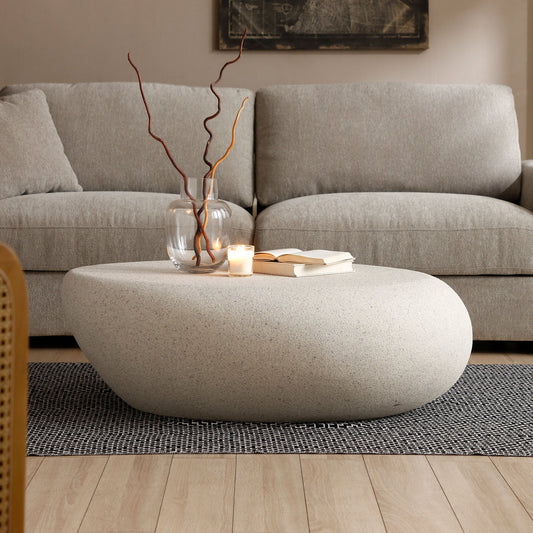 A beige elliptical coffee table in a modern living room. A vase with branches and a lit candle are on the table, with an open book nearby.