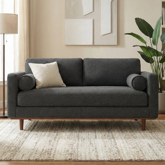 A gray sofa with rolled armrests and a white textured pillow, placed on a beige rug in a well-lit room with framed artwork on the wall and a potted plant to the side