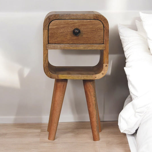 A solid wood bedside table with an open drawer and curved design, placed next to a bed with white pillows.