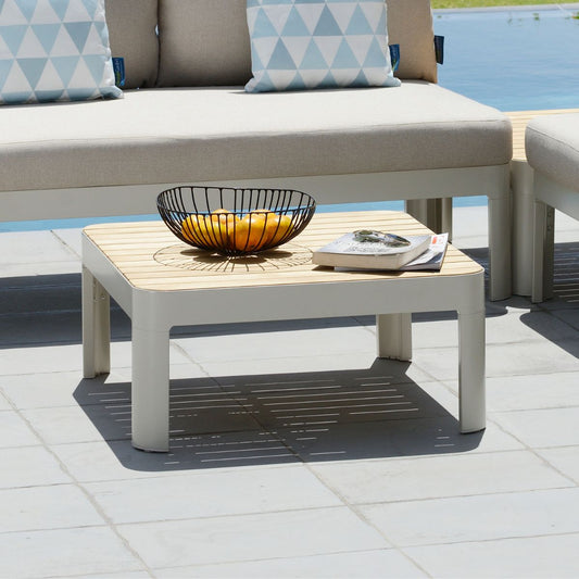 A light brown wooden top coffee table with a white frame, topped with a wire basket of oranges, situated near a poolside lounge sofa setup.