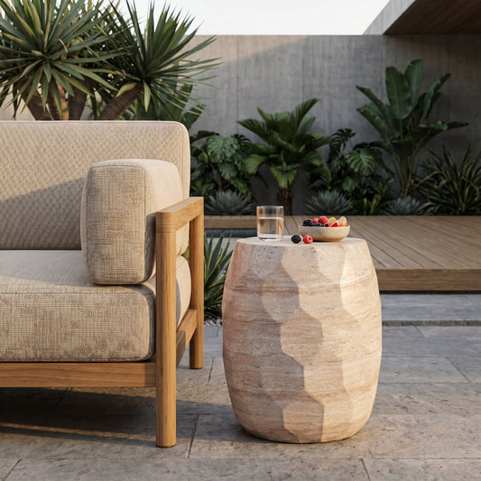 Sleek outdoor concrete side table with faceted drum design, placed on a stone patio beside a sofa and surrounded by green plants.