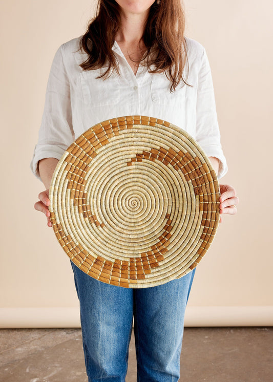 A person holding a circular wall basket with a radial pattern of light and dark natural wood tones.