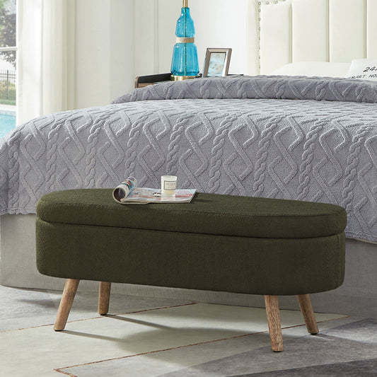 Olive green oval storage ottoman with wooden legs, set in a bedroom with grey bedding and decor items.