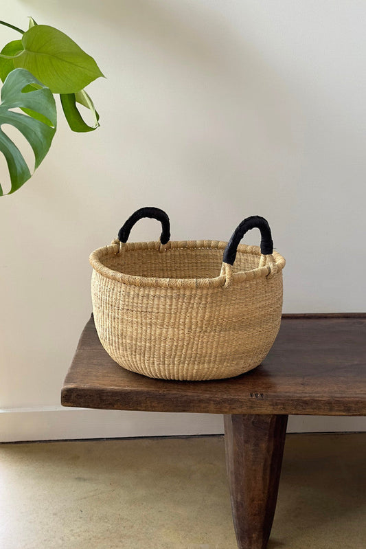 Natural woven basket with black handles resting on wooden table with green leaf in soft light
