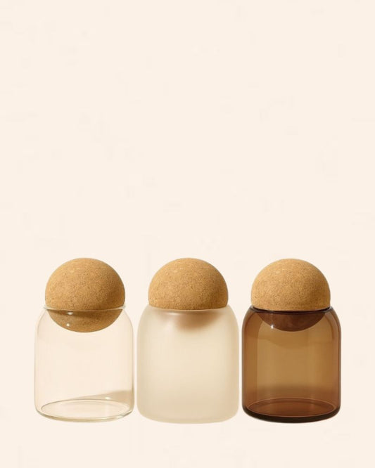Three glass jars with cork lids in clear, beige, and amber colors.