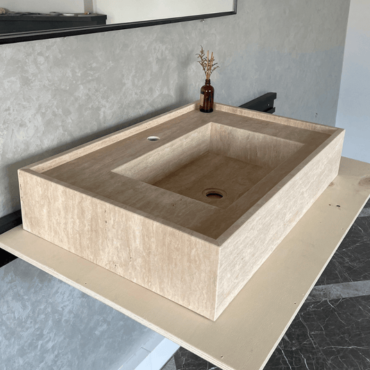 A rectangular beige handcrafted travertine sink basin placed on a modern countertop, with a decorative terrarium of dried herbs in the background.