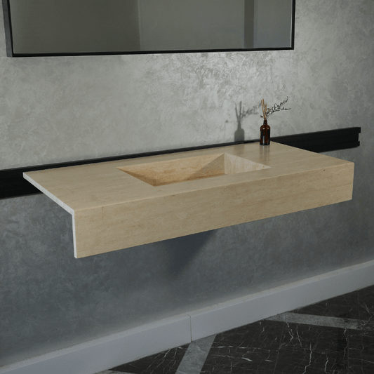 A rectangular handcrafted travertine sink with a natural stone finish is displayed on a wall, with a dark mirror above it and a small vase of plants beside it, situated in a room with a gray textured wall and black-and-white tiled floor.